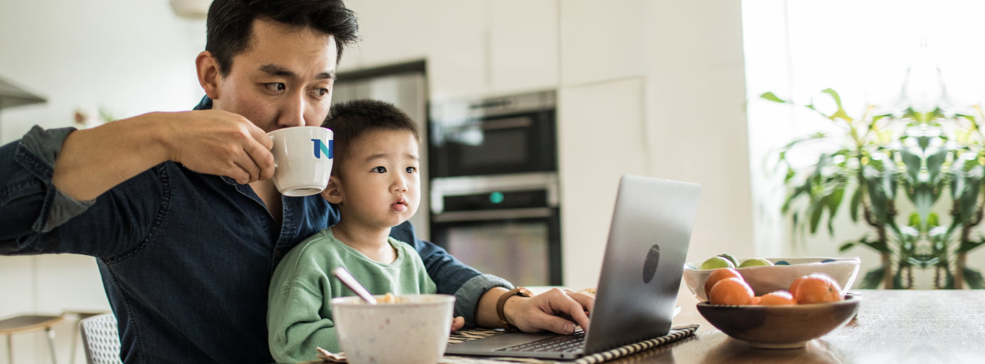 father working on laptop at kitchen table sipping from NU mug with toddler son in his lab